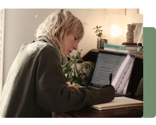 Jo Delacourt working at a desk, using a laptop and taking notes in a notebook, surrounded by plants and books in soft natural light.