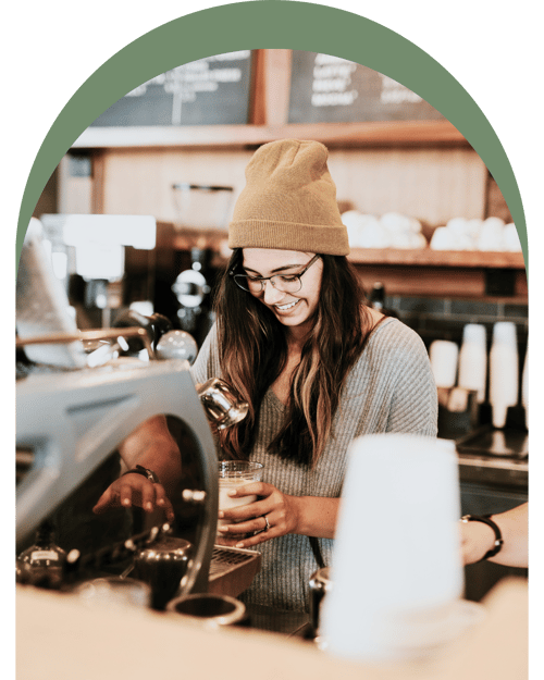 A barista in a cozy café, wearing a yellow beanie, prepares a drink behind a coffee machine, with pastries on display above.