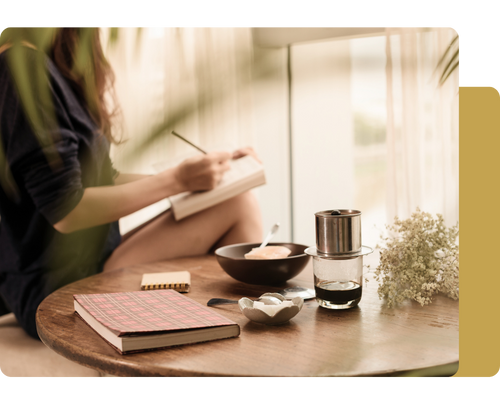 A person writes in a notebook beside a bowl of food, coffee, and flowers on a wooden table, bathed in soft natural light.
