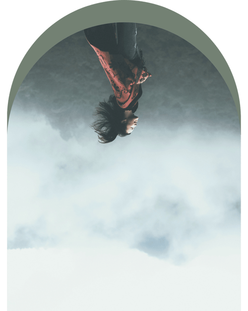 A person stands in front of a misty landscape, wearing a vibrant red and black garment, with hair billowing in the breeze.