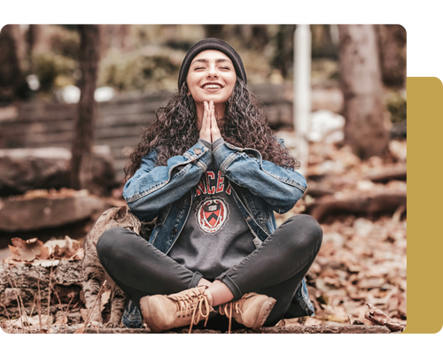 A person sits cross-legged on a forest floor covered in leaves, wearing a denim jacket and boots, with hands pressed in a prayer position. A person sits cross-legged on a forest floor covered in leaves, wearing a denim jacket and boots, with hands pressed in a prayer position.