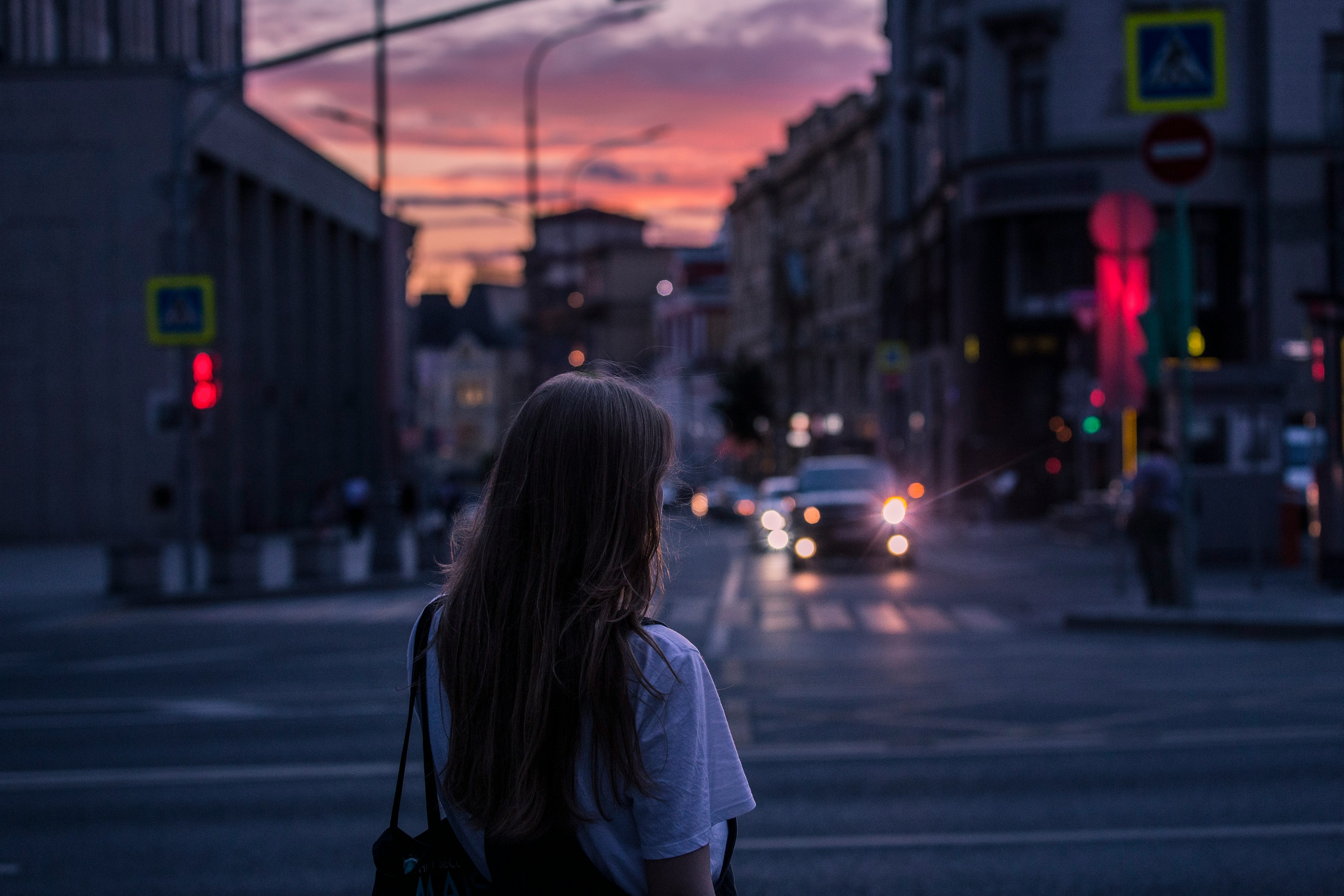 A woman with long hair stands on a city street at dusk, watching oncoming traffic and a colorful sunset in the background.