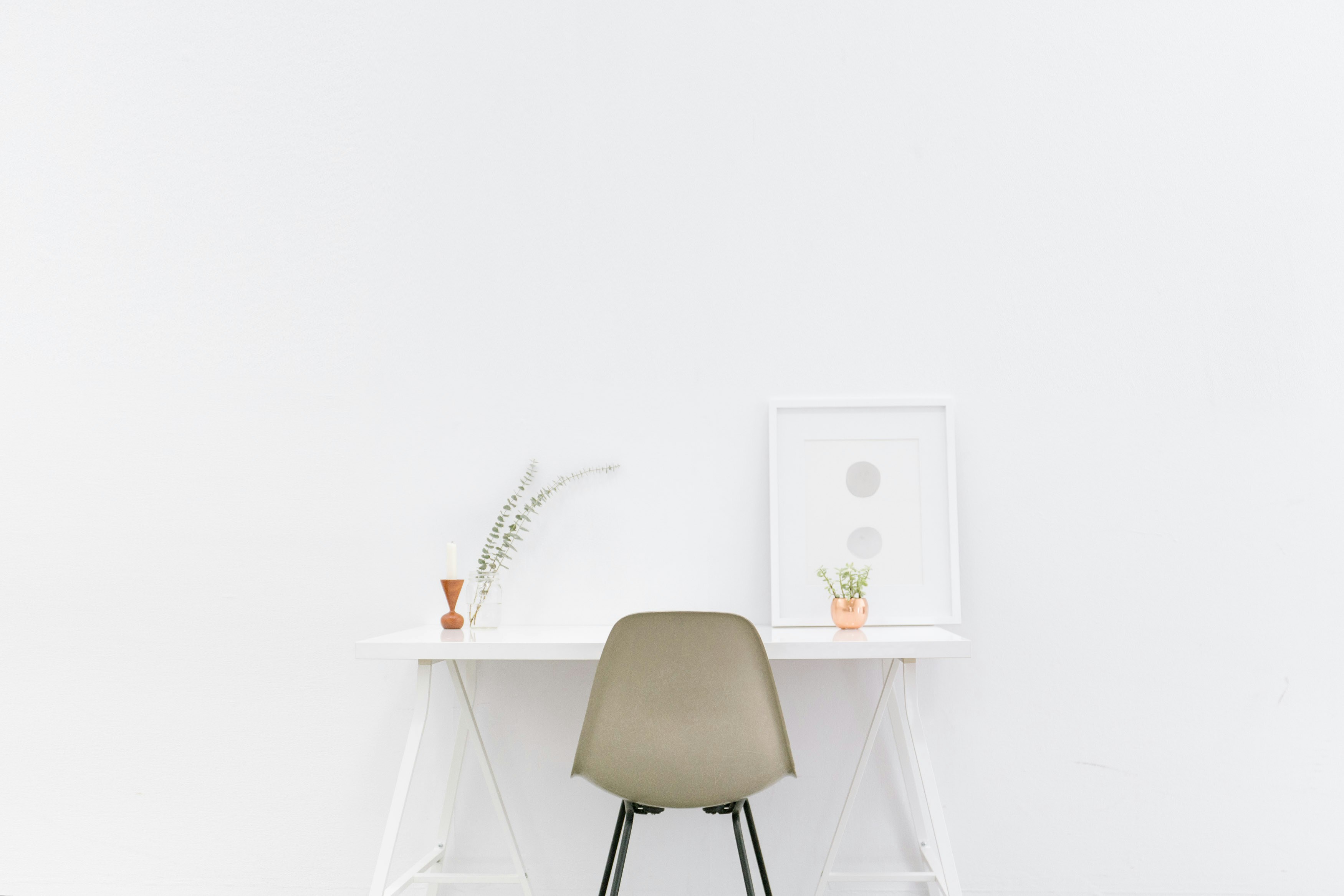 Minimalist workspace with a white desk, an olive green chair, and decorative elements including vases and a framed print on a white wall.