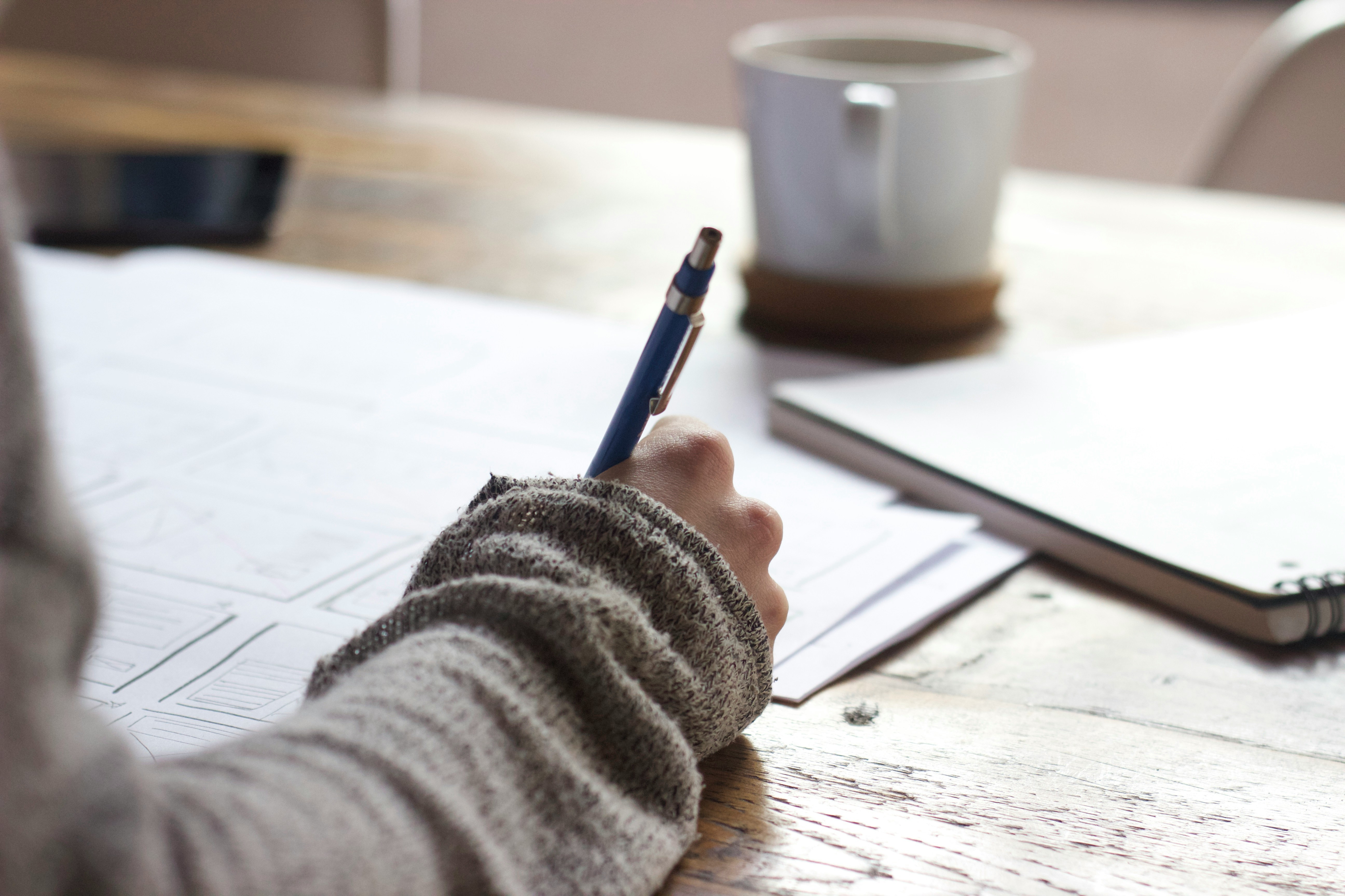 A person in a gray sweater writes notes with a blue pen on sketch papers, a coffee cup, and a notebook nearby on a wooden table.
